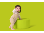 Baby im beigen Overall steht neben einem grünen Pouf und schaut in die Kamera.