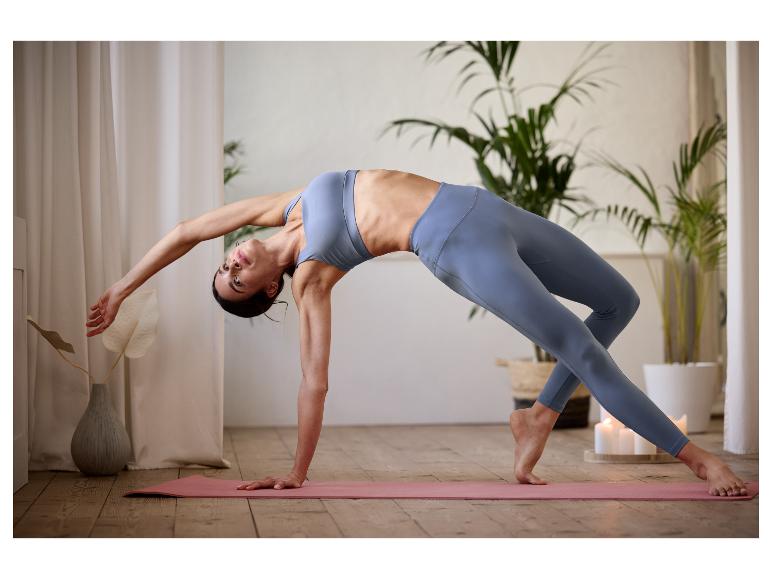 Eine Frau in einem blauen Sport-BH und Leggings macht Yoga auf einer rosafarbenen Matte.