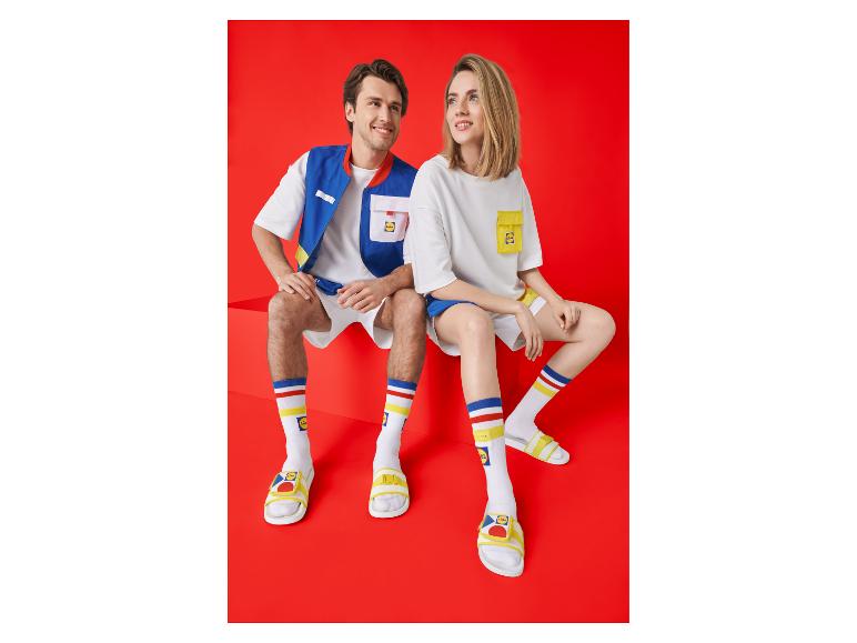Zwei Personen in bunten T-Shirts, Westen, Shorts, Socken und Sandalen.