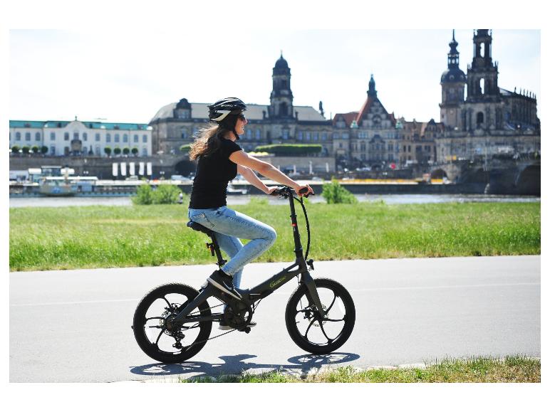 Frau fährt Sachsengrad E-Bike auf einem Flussweg mit Stadtansicht im Hintergrund.