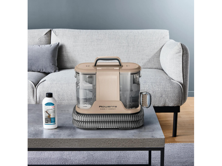 Beiger kompakter Teppichreiniger auf einem Couchtisch mit Reinigungslösung und grauem Sofa.