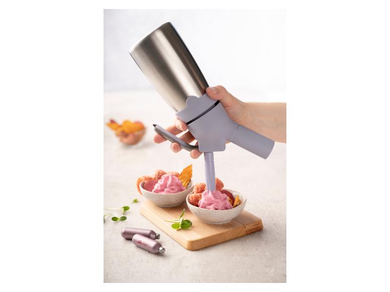 Eine Hand verwendet einen Küchensyphon, um rosa Schaum in Schalen mit Trockenfrüchten zu füllen.