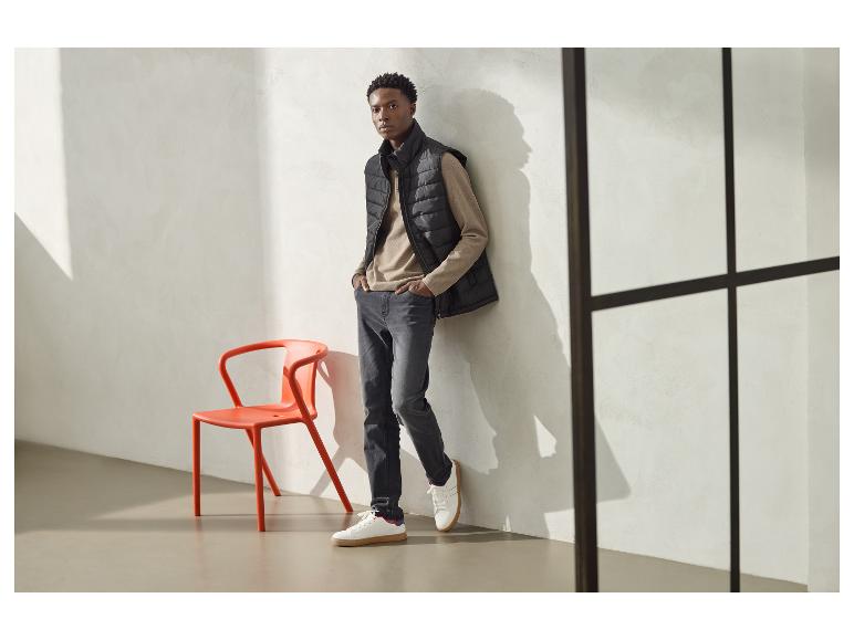 Mann in schwarzer Steppweste, beigem Langarmshirt und grauer Jeans, an eine Wand gelehnt.