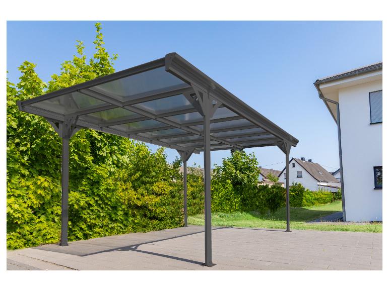 Graues Carport mit transparentem Dach auf gepflasterter Einfahrt neben grüner Hecke und Haus.