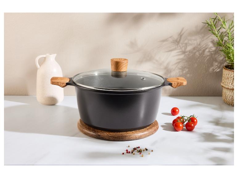 Schwarzer Topf mit Holzgriffen und Glasdeckel auf einem Holzuntersetzer, mit Tomaten und Gewürzen.