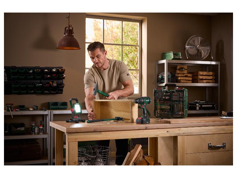 Mann montiert eine Holzkiste mit einem Hammer, umgeben von Elektrowerkzeugen und Werkstattausrüstung.