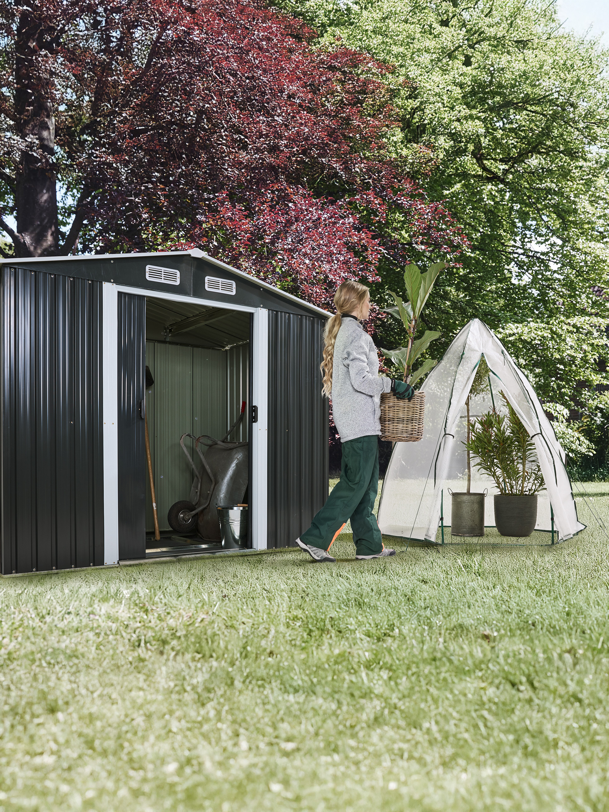 Frau trägt Pflanze in einem Korb, neben einem Gartenhaus und einem kleinen Gewächshaus.