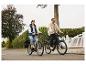 Zwei Personen fahren E-Bikes auf einer Straße, eine Frau auf einem hellen und ein Mann auf einem dunklen Rad.