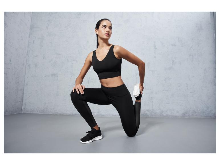 Frau in schwarzem Sport-BH, Leggings und Sneakers dehnt sich vor einem grauen Hintergrund.