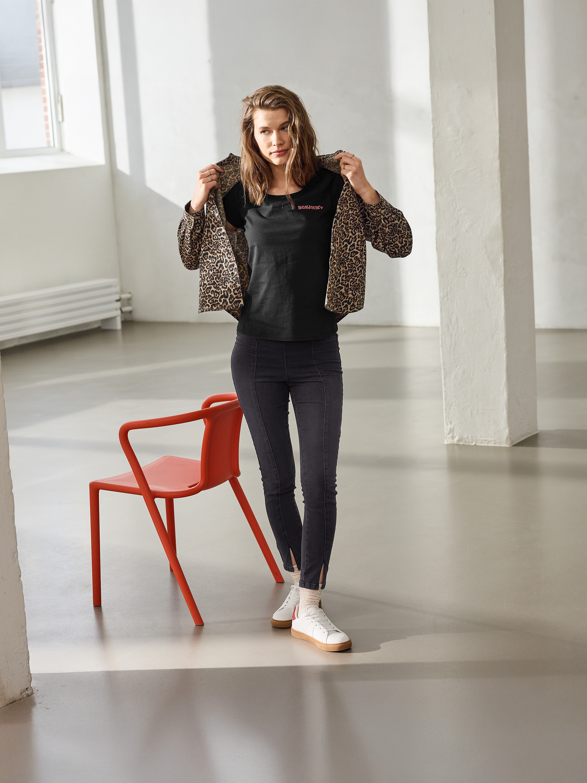 Frau in Leopardenjacke, schwarzem 'BONJOUR' T-Shirt und schwarzer Skinny Jeans, posiert neben einem roten Stuhl.