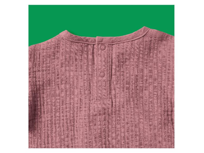 Rosa Musselinbluse mit Knöpfen im Nacken, vor grünem Hintergrund.