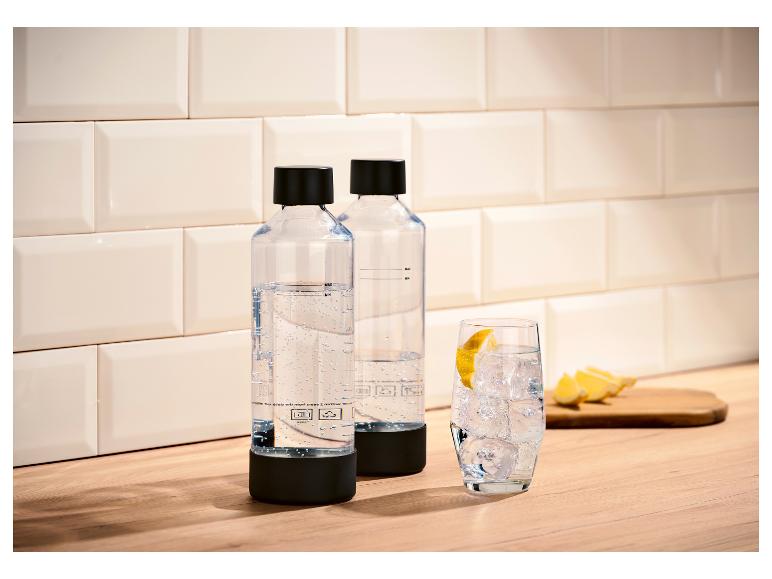 Zwei klare Flaschen Sprudelwasser mit schwarzen Verschlüssen auf einer Holztheke, neben einem Glas Eiswasser mit Zitrone.