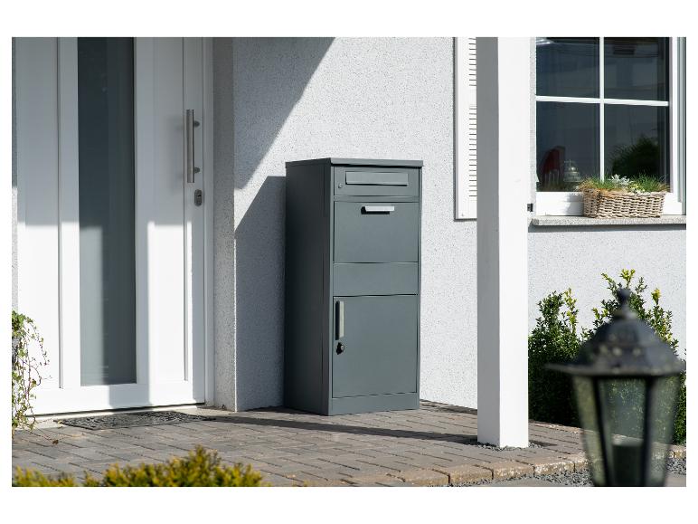 Dunkelgrauer Paket- und Briefkasten neben einer weißen Haustür auf einer gepflasterten Terrasse.