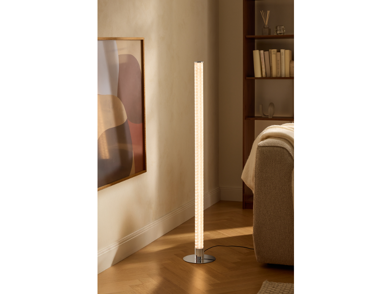 Zylindrische Stehlampe mit Chromfuß beleuchtet ein modernes Wohnzimmer mit Bücherregal und Sofa.