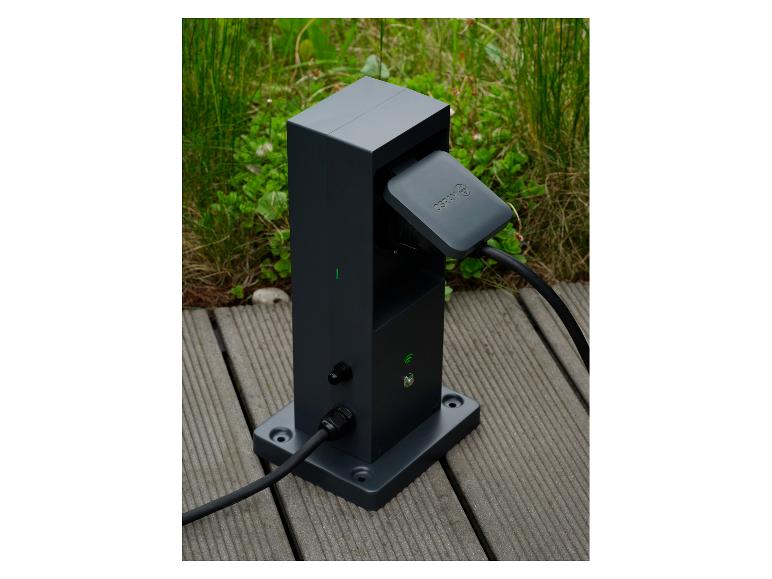 Dunkelgraue Ladestation für Elektrofahrzeuge mit angeschlossenem Kabel auf einer Holzterrasse.