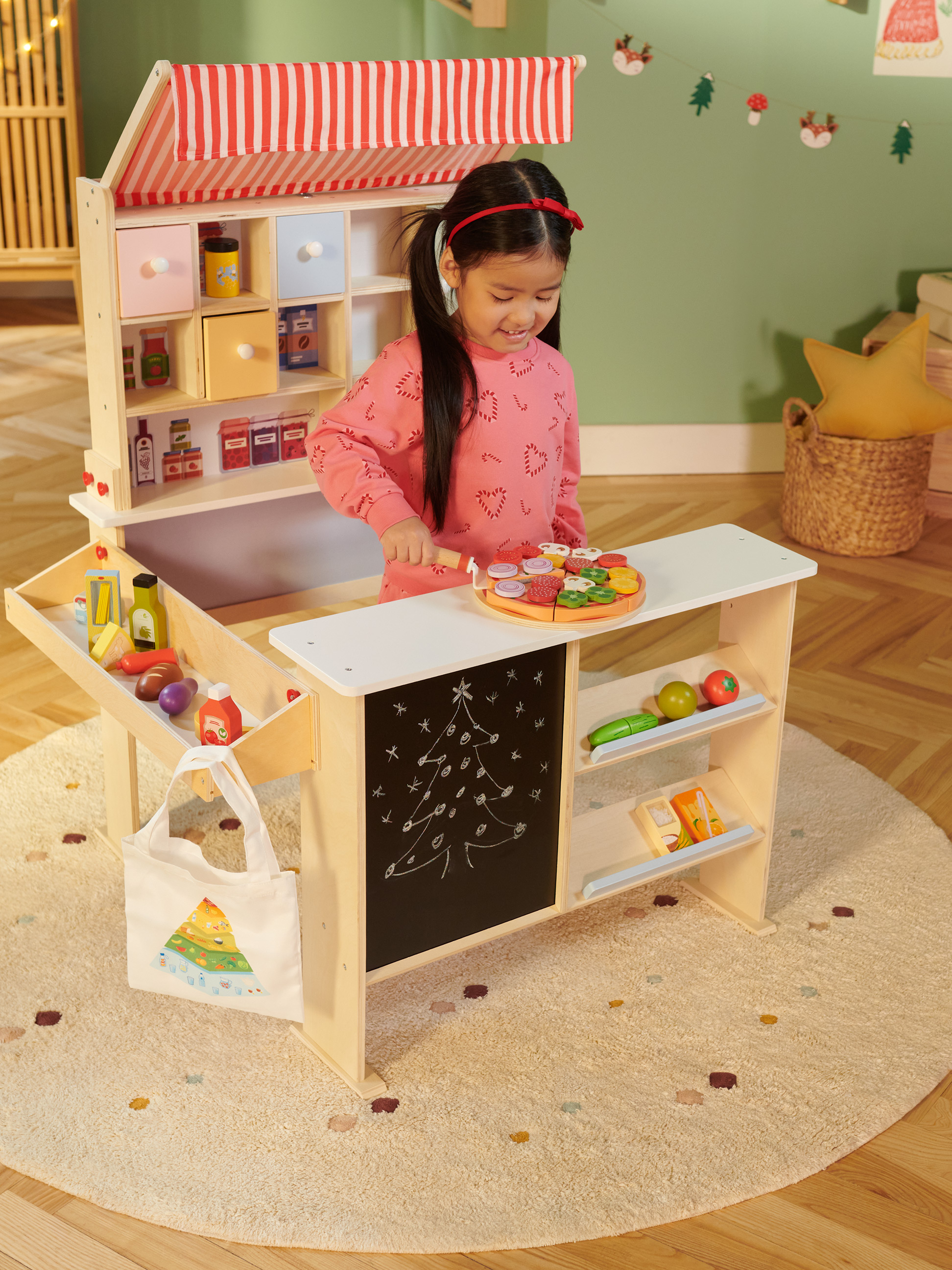 Lächelndes Mädchen spielt mit einem Holz-Kaufladen, der mit Spielzeugessen und einer Kreidetafel ausgestattet ist.