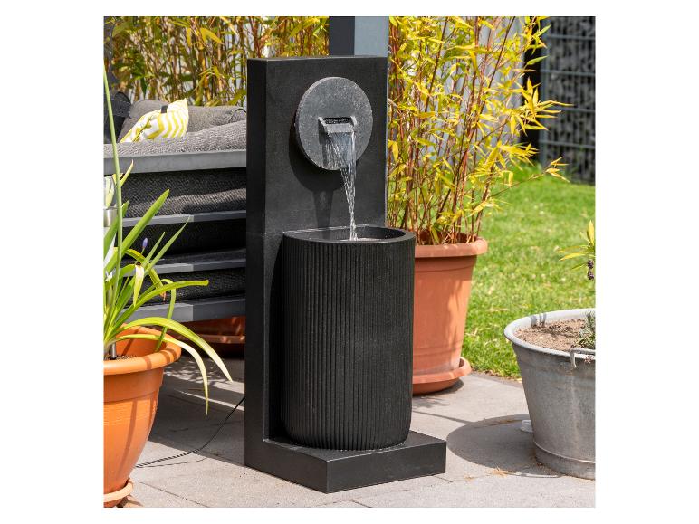 Moderner schwarzer Gartenbrunnen mit Wasser, das in ein geriffeltes Becken fließt, umgeben von Topfpflanzen.