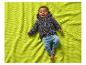 Ein lächelndes Baby in grauem Cardigan und blauer Hose, liegend auf einer grünen Decke.