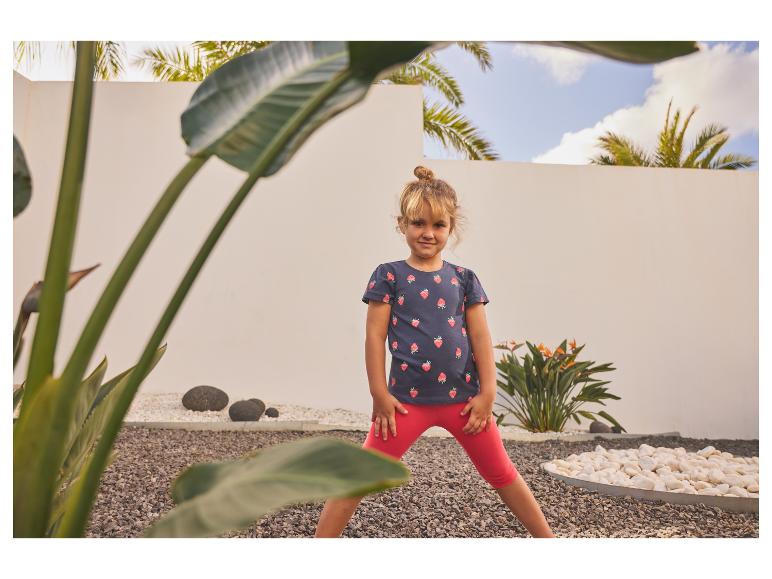 Mädchen in Erdbeer-T-Shirt und roten Leggings, steht in einem Garten.