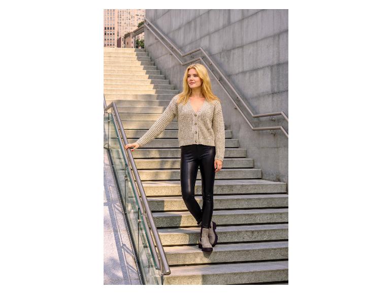Frau in beiger Strickjacke und schwarzen Kunstleder-Leggings auf Betontreppe.