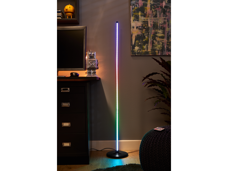 Eine hohe, farbwechselnde LED-Stehlampe steht neben einem dunklen Schreibtisch mit Computermonitor.