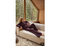 Frau in pflaumenfarbener Loungewear entspannt auf einem Sofa an einem großen Fenster mit Waldblick.
