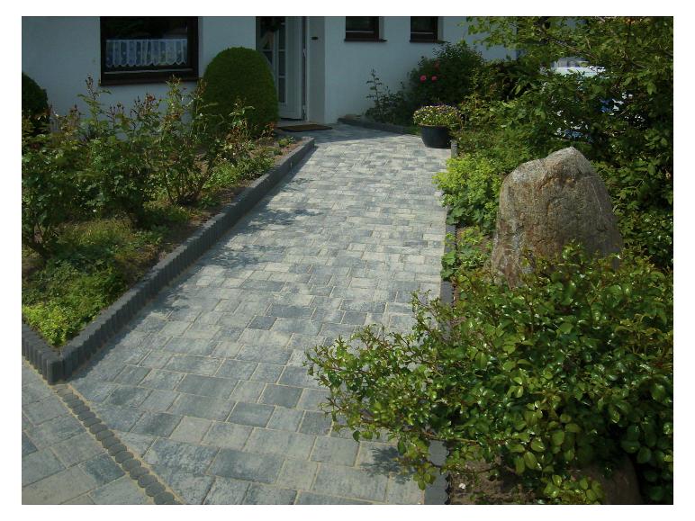 Gepflasterter Weg führt zu einem Haus, gesäumt von Blumenbeeten und Grün.