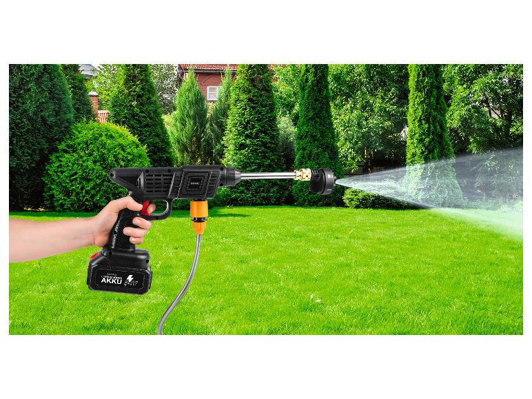 Akku-Hochdruckreiniger in der Hand, spritzt Wasser auf grünen Rasen mit Sträuchern und Haus im Hintergrund.
