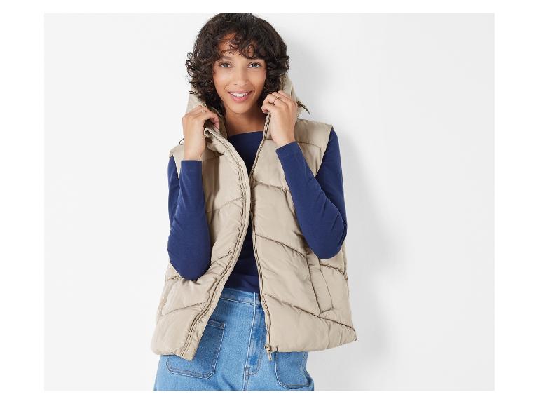 Eine Frau trägt eine beige Weste, ein blaues Langarmshirt und Jeans.