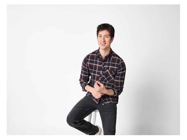 Lächelnder Mann in kariertem Hemd und Jeans, auf einem Hocker sitzend.