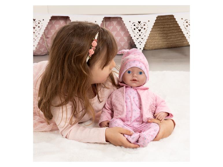 Mädchen spielt mit einer Babypuppe in rosa Kleidung auf einem weißen Teppich.