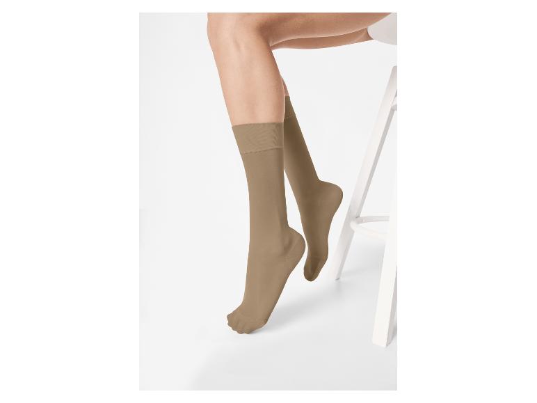Frauenbeine in hautfarbenen Socken, auf einem weißen Hocker sitzend.