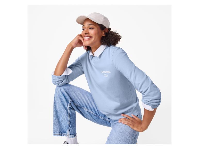 Frau in hellblauem Sweatshirt mit „Breakfast Club“-Aufschrift, Jeans und Kappe.