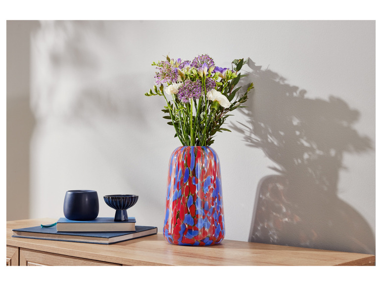 Blumen in einer roten und blauen Vase mit Punkten, zwei blaue Dekoartikel und Bücher auf einem Holztisch.