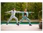 Zwei Personen in Trainingsanzügen und Sportschuhen machen Yoga im Freien auf einer Holzterrasse.