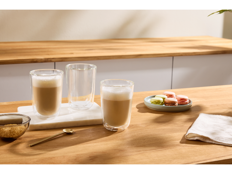 Zwei Gläser Kaffee Latte, ein leeres Glas, Macarons und brauner Zucker auf einem Holztisch.