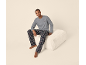 Mann in grauem Langarmshirt mit „Happy Holidays“-Aufschrift und karierter Pyjamahose, sitzt auf einem Hocker.