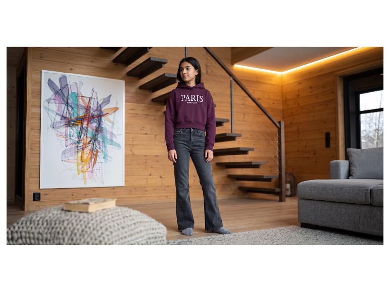 Mädchen in einem bordeauxroten Hoodie mit „PARIS AMOUR CLUB“-Aufschrift und dunklen Jeans, stehend in einem modernen Wohnzimmer.