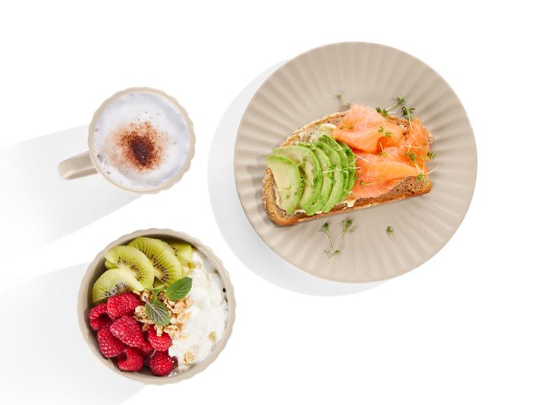 Ein Teller mit geräuchertem Lachs auf Toast, Avocado, eine Schüssel mit Quark, Kiwi und Himbeeren und eine Tasse Cappuccino.