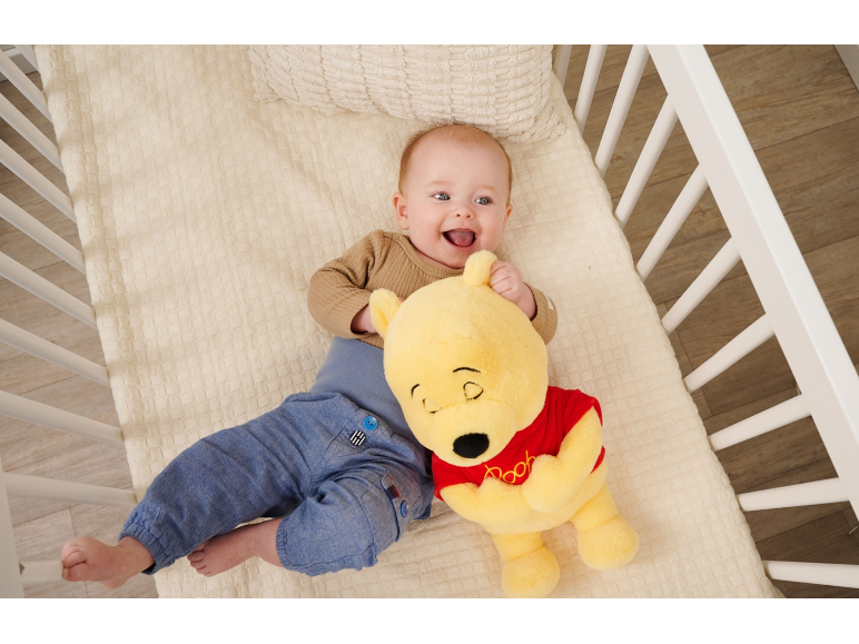 Lächelndes Baby liegt mit einem Plüschbären im Gitterbett.