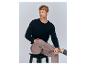 Mann in schwarzem Pullover und beiger Hose sitzt auf einem Hocker.