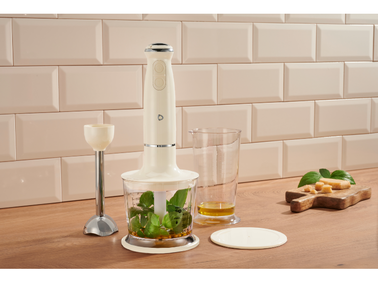 Cremefarbener Stabmixer mit Kräutern, Öl und Käse auf einer Holzoberfläche.