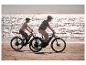 Zwei Personen auf E-Bikes fahren an einem Sandstrand mit glitzerndem Wasser im Hintergrund.