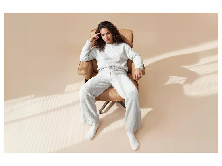 Frau in gemütlicher weißer Loungewear, sitzt in einem braunen Sessel mit Fensterlicht.