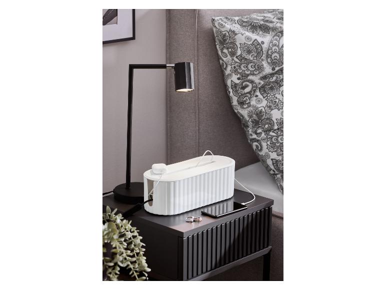 Schwarzer Nachttisch mit Tischlampe, weißer Kabelbox und aufladendem Smartphone.