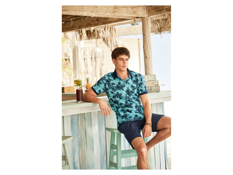 Mann in einem blauen Polohemd mit Palmenprint und blauen Shorts, der auf einer Bar sitzt.