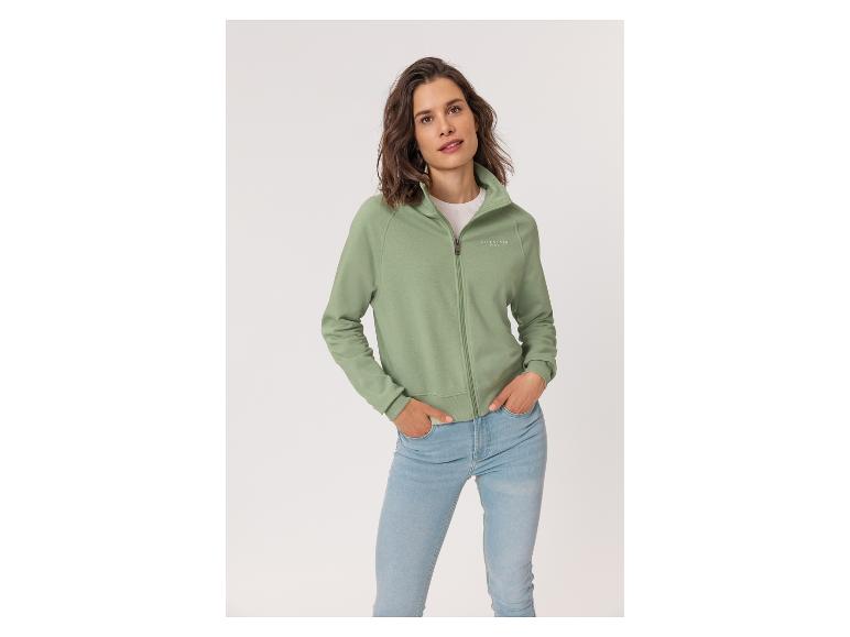 Frau in einer grünen Strickjacke mit Reißverschluss und Jeans, Hände in den Taschen.