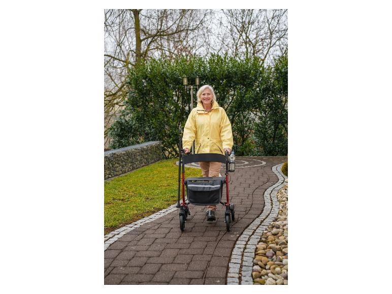 Ältere Frau in gelber Jacke geht mit Rollator auf gepflastertem Weg.