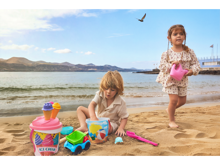 Zwei Kinder spielen mit Sandspielzeug, darunter ein Eiskübel und eine Gießkanne, am Strand.