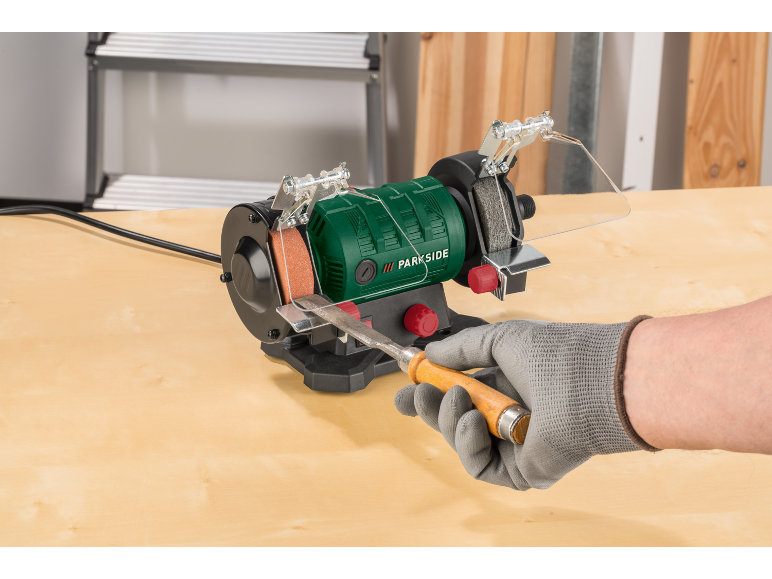 Grüner Doppelschleifbock mit einem Holzmeißel, der von einer behandschuhten Hand geschliffen wird.